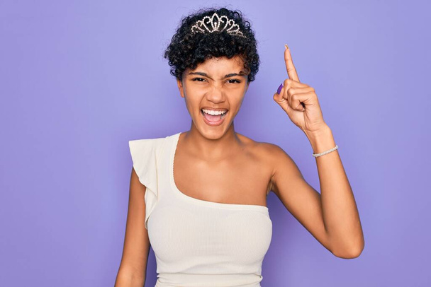 A fiatal, gyönyörű afro-amerikai nő tiara koronát visel a lila háttér felett, ujjal mutogatva a sikeres ötlettel. Kilépett és boldog. Egyes számú.. - Fotó, kép