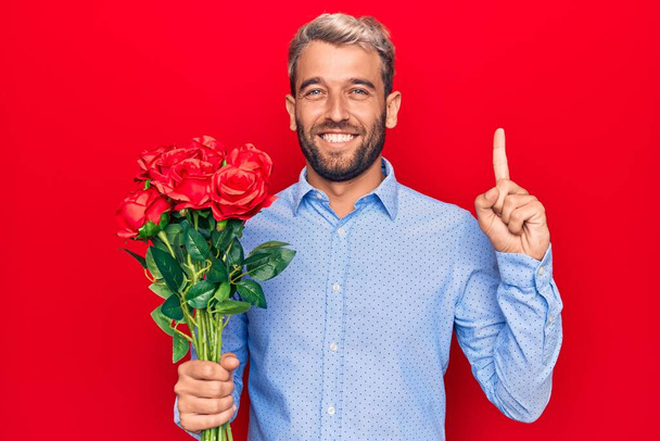 Молодой красивый блондин романтик с букетом красивых роз на красном фоне улыбается с идеей или вопросом указывая пальцем со счастливым лицом, номер один
 - Фото, изображение
