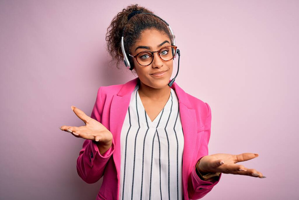 ヘッドセットを使用して作業眼鏡を着用して若いアフリカ系アメリカ人のコールセンターエージェントの女の子は、腕や手を上げと混乱表現。疑いの概念. - 写真・画像