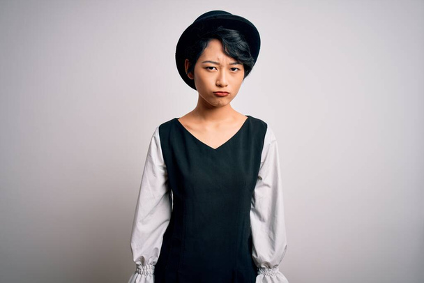 Jong mooi aziatisch meisje dragen casual jurk en hoed staan over geïsoleerde witte achtergrond depressief en zorgen te maken voor verdriet, huilen boos en bang. Droevige uitdrukking. - Foto, afbeelding