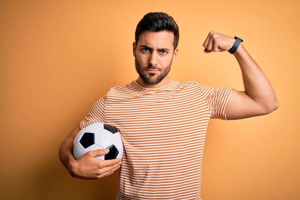 Красивый игрок с бородой играет в футбол держа футбольный мяч на желтом фоне сильный человек показывает мышцы рук, уверенный и гордый властью
 - Фото, изображение