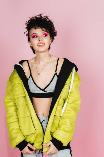 Beautiful teenager girl sincerely smiles and laughs in sports stylish clothes in the studio on a pink background. Bright girl with a short flying hairstyle in a puffed neon lemon jacket and pink smokey eyes. - 写真・画像