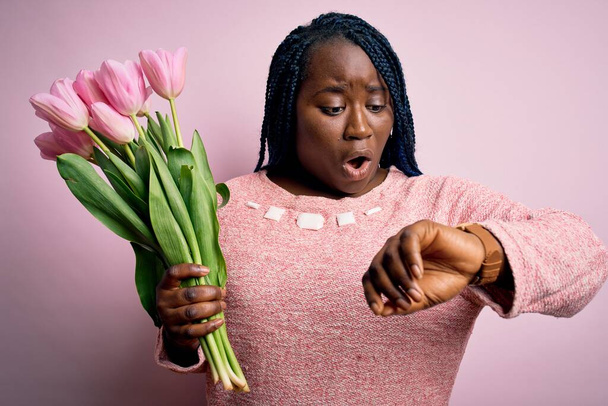 Jovem afro-americana plus size mulher com tranças segurando buquê de tulipas rosa flor Olhando para o relógio tempo preocupado, com medo de se atrasar
 - Foto, Imagem