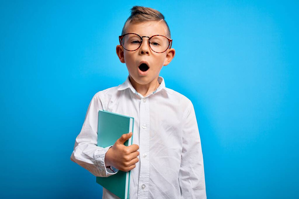 Jeune enfant étudiant caucasien portant des lunettes intelligentes et tenant un livre de l'école effrayé en état de choc avec un visage surprise, effrayé et excité par l'expression de la peur - Photo, image