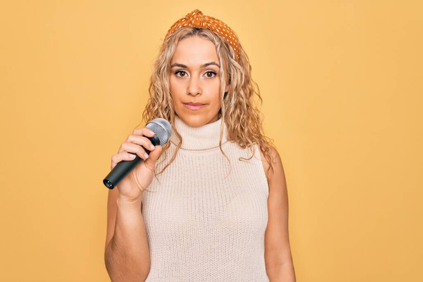 Jonge mooie blonde zangeres zingen met behulp van microfoon over gele achtergrond denken houding en sobere expressie op zoek zelfverzekerd - Foto, afbeelding