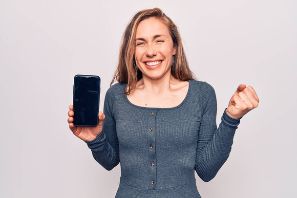 Young beautiful blonde woman showing screen of smartphone screaming proud, celebrating victory and success very excited with raised arm  - Фото, изображение