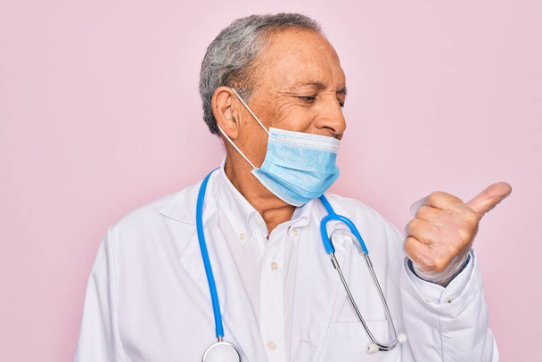 Senior hoary doctor man wearing medical mask and stethoscope over pink background pointing and showing with thumb up to the side with happy face smiling - Foto, afbeelding