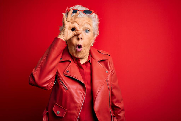 Senior beautiful grey-haired woman wearing casual red jacket and sunglasses doing ok gesture shocked with surprised face, eye looking through fingers. Unbelieving expression. - Zdjęcie, obraz