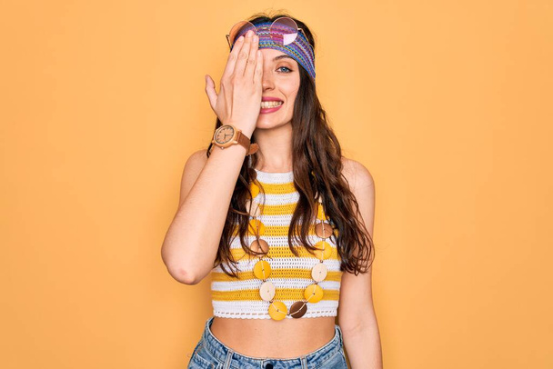 Young beautiful hippie woman with blue eyes wearing accesories and sunnglasses covering one eye with hand, confident smile on face and surprise emotion. - Photo, Image