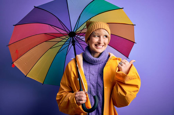 Mladá blondýnka s krátkými vlasy nosí nepromokavý plášť a barevný deštník ukazuje palcem nahoru na stranu se šťastnou tváří usmívá - Fotografie, Obrázek