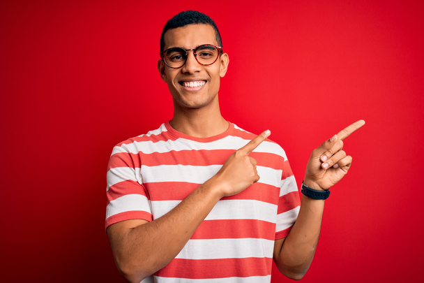 Jovem afro-americano bonito vestindo camiseta listrada casual e óculos sorrindo e olhando para a câmera apontando com duas mãos e dedos para o lado
. - Foto, Imagem