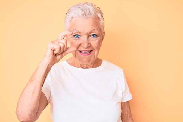 Senior gyönyörű nő kék szemek és szürke haj visel klasszikus fehér póló sárga háttér mosolygós és magabiztos gesztikulálás kézzel csinál kis méretű jel ujjak keres, és a kamera. intézkedés koncepciója.  - Fotó, kép