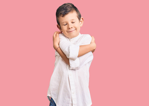 エレガントなシャツを着てかわいいブロンドの子供は、自分自身が幸せと肯定的な抱擁、自信を笑顔。自己愛と自己ケア  - 写真・画像
