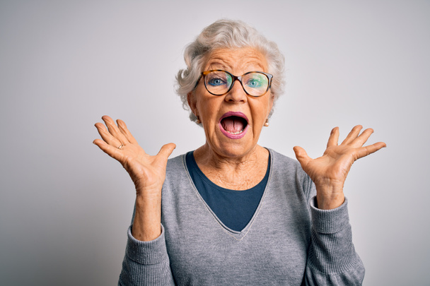 Senior krásná šedovlasá žena na sobě ležérní svetr a brýle na bílém pozadí slaví šílený a ohromen pro úspěch s rukama zvednutýma a otevřené oči křičí vzrušeně. Koncept vítěze - Fotografie, Obrázek