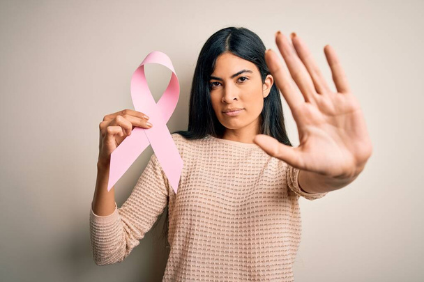 Joven mujer hispana hermosa sosteniendo la conciencia del cáncer de mama cinta rosa con la mano abierta haciendo señal de stop con expresión seria y segura, gesto de defensa
 - Foto, imagen