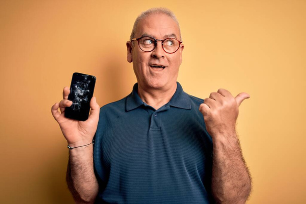 Middle age man holding broken smartphone showing cracked screen over yellow background pointing and showing with thumb up to the side with happy face smiling - Fotó, kép