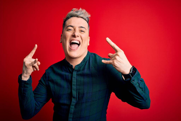 Young handsome modern man wearing elegant green shirt over red isolated background shouting with crazy expression doing rock symbol with hands up. Music star. Heavy music concept. - Photo, Image