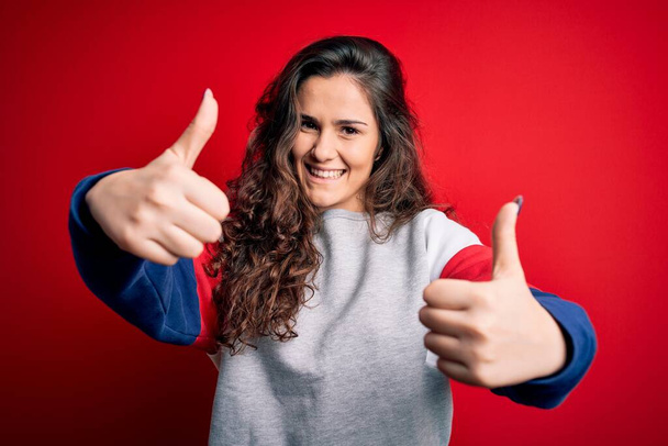 Jonge mooie vrouw met krullend haar dragen casual sweatshirt over geïsoleerde rode achtergrond goed te keuren doen positieve gebaar met de hand, duimen omhoog glimlachend en blij voor succes. Winnaar gebaar. - Foto, afbeelding