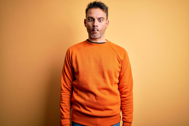 Young handsome man wearing orange casual sweater standing over isolated yellow background making fish face with lips, crazy and comical gesture. Funny expression. - Photo, image