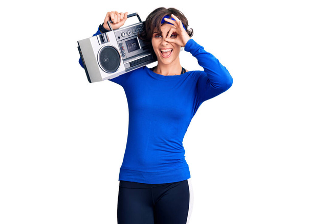 Beautiful young woman with short hair wearing workout clothes and holding boombox smiling happy doing ok sign with hand on eye looking through fingers  - Photo, Image
