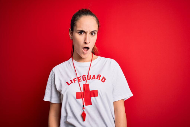 Όμορφη ναυαγοσώστρια γυναίκα που φοράει μπλουζάκι με κόκκινο σταυρό χρησιμοποιώντας σφυρίχτρα πάνω σε απομονωμένο φόντο στο σοκαριστικό πρόσωπο, δείχνοντας σκεπτική και σαρκαστική, έκπληκτη με ανοιχτό στόμα - Φωτογραφία, εικόνα