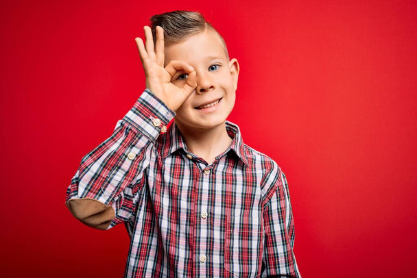 Young little caucasian kid with blue eyes standing wearing elegant shirt over red background doing ok gesture with hand smiling, eye looking through fingers with happy face. - Photo, Image