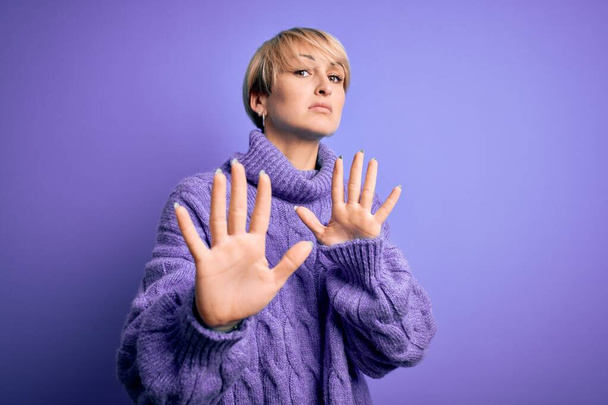 Mujer rubia joven con el pelo corto con suéter de cuello alto de invierno sobre fondo púrpura Alejando las palmas de las manos que muestran rechazo y negación con expresión de miedo y repugnante. Detente y prohibido
. - Foto, imagen