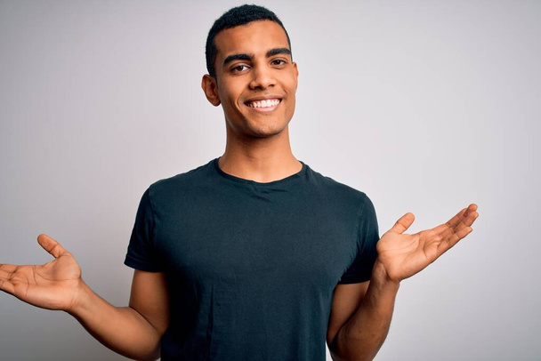 Junger gutaussehender afrikanisch-amerikanischer Mann in lässigem T-Shirt vor weißem Hintergrund lächelnd mit beiden Händen geöffneten Handflächen, Präsentation und Werbung Vergleich und Ausgewogenheit - Foto, Bild