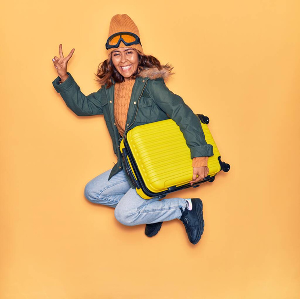 Νεαρή όμορφη Λατίνα γυναίκα που φοράει χειμωνιάτικα ρούχα χαμογελώντας ευτυχισμένη. Άλμα με χαμόγελο στο πρόσωπο κρατώντας τσάντα καμπίνα κάνει σήμα νίκη πάνω από απομονωμένο κίτρινο φόντο - Φωτογραφία, εικόνα