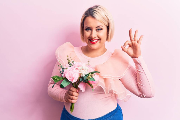 Linda loira plus size mulher segurando buquê de flores cor de rosa sobre fundo isolado fazendo sinal ok com os dedos, sorrindo amigável gesto excelente símbolo
 - Foto, Imagem