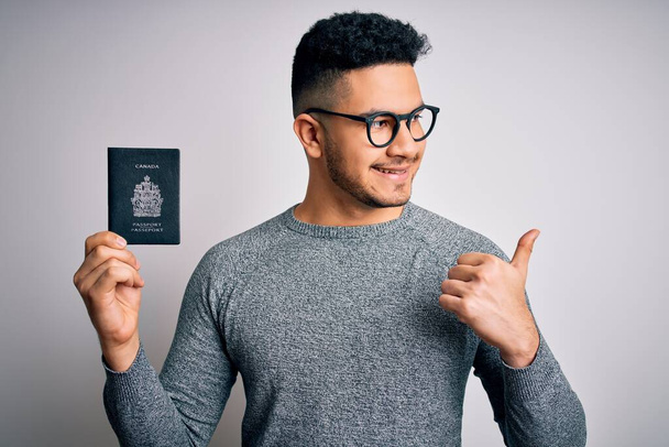 Молодий гарний турист у відпустці в окулярах тримає канадський паспорт вказуючи пальцем до сторони з щасливим обличчям посміхаючись - Фото, зображення