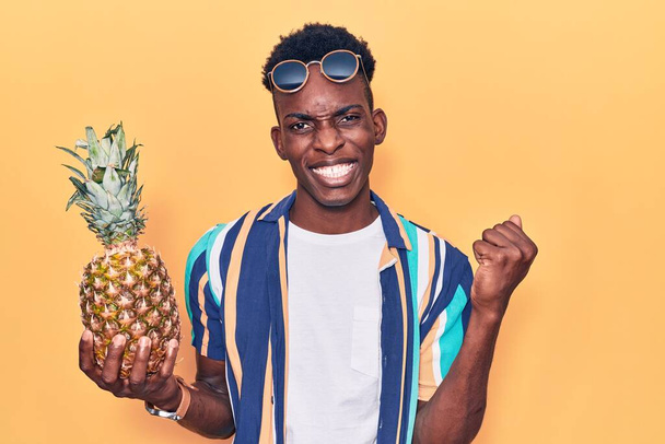 Jonge Afro-Amerikaanse man draagt zomerkleren en een zonnebril met ananas die trots schreeuwt en de overwinning en het succes viert.  - Foto, afbeelding
