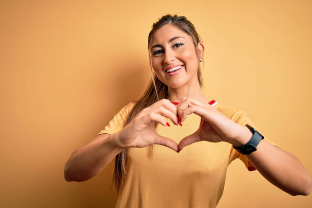 Молода красива атлетична жінка в повсякденній футболці і хвіст на жовтому тлі посміхається в коханні, роблячи символ серця фігурою рук. Романтична концепція
. - Фото, зображення