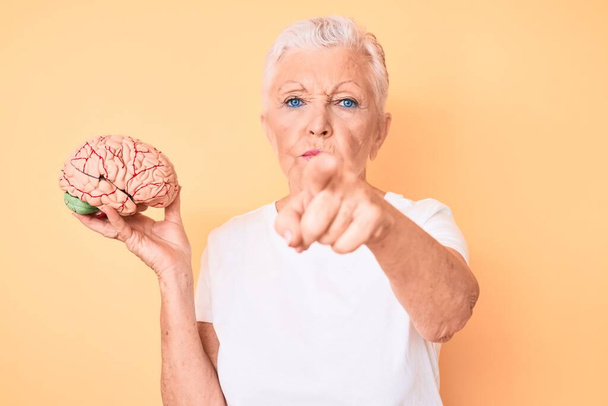 Senior krásná žena s modrýma očima a šedé vlasy drží mozek jako koncept duševního zdraví ukazuje prstem na fotoaparát a na vás, sebevědomé gesto vypadá vážně  - Fotografie, Obrázek