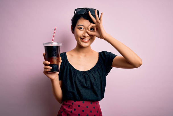 Junge schöne chinesische Frau trinkt Cola sprudelnden Getränk mit Stroh über rosa Hintergrund mit glücklichem Gesicht lächelnd ok Zeichen mit Hand auf Auge Blick durch Finger - Foto, Bild