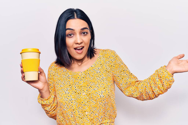 Junge schöne lateinische Frau mit einer Tasse Kaffee zum Mitnehmen feiert Leistung mit glücklichem Lächeln und Siegerausdruck mit erhobener Hand  - Foto, Bild