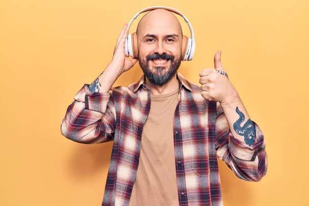 Jovem homem bonito ouvindo música usando fones de ouvido sorrindo feliz e positivo, polegar para cima fazendo excelente e sinal de aprovação  - Foto, Imagem