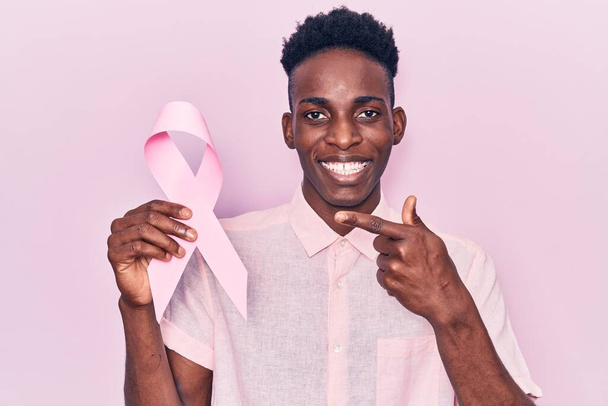 Młody, afrykański Amerykanin trzymający różową wstążkę nowotworową uśmiechnięty szczęśliwy wskazując dłonią i palcem  - Zdjęcie, obraz