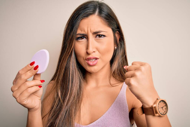 Young beautiful woman using beauty sponge with facial makeup over pink background annoyed and frustrated shouting with anger, crazy and yelling with raised hand, anger concept - Photo, Image