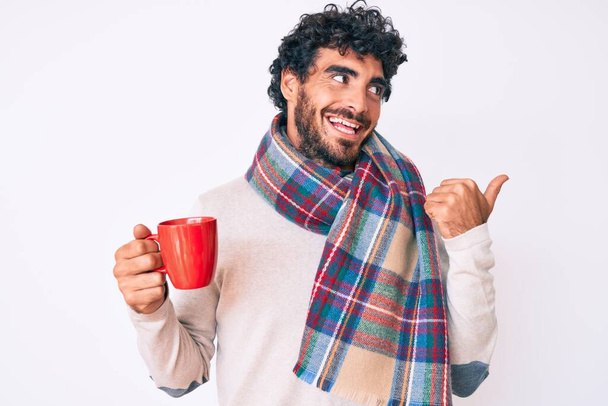 Przystojny młody człowiek z kręconymi włosami i niedźwiedź noszący zimowy szalik i pijący filiżankę gorącej kawy z kciukiem w górze, uśmiechnięty szczęśliwy z otwartymi ustami.  - Zdjęcie, obraz