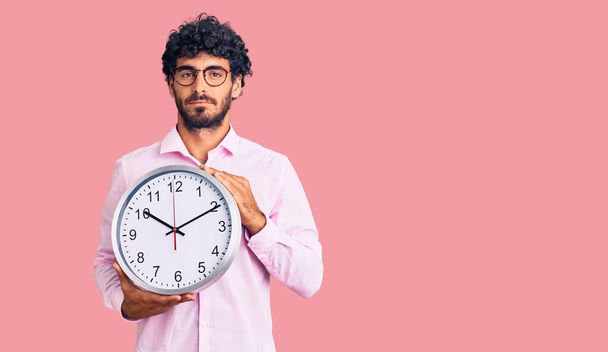Handsome young man with curly hair and bear holding big clock thinking attitude and sober expression looking self confident  - Photo, Image