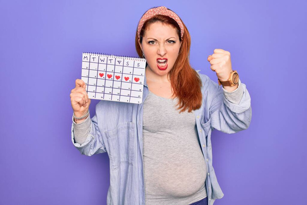 Junge schöne rothaarige schwangere Frau erwartet Baby Wartezeit Kalender genervt und frustriert schreien vor Wut, verrückt und schreien mit erhobener Hand, Wut Konzept - Foto, Bild