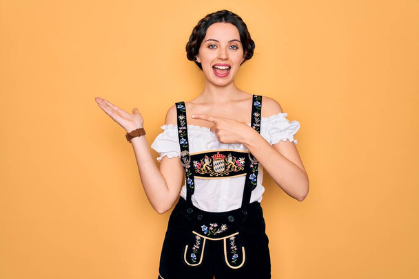Young beautiful german woman with blue eyes wearing traditional octoberfest dress amazed and smiling to the camera while presenting with hand and pointing with finger. - Photo, Image