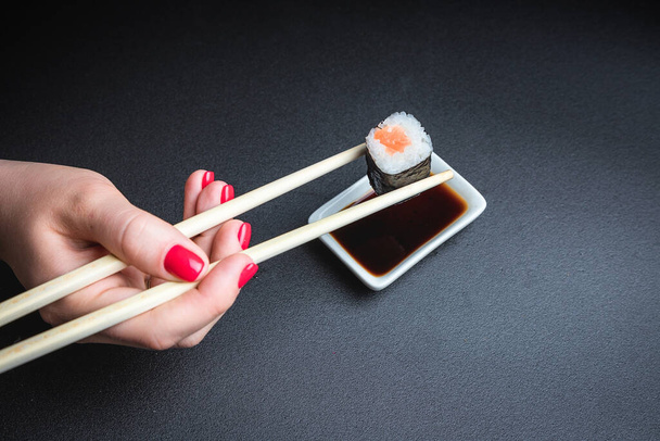 A hand holds a sushi roll with chopsticks, on a black background. Oriental cuisine. Japanese culture. - Foto, afbeelding