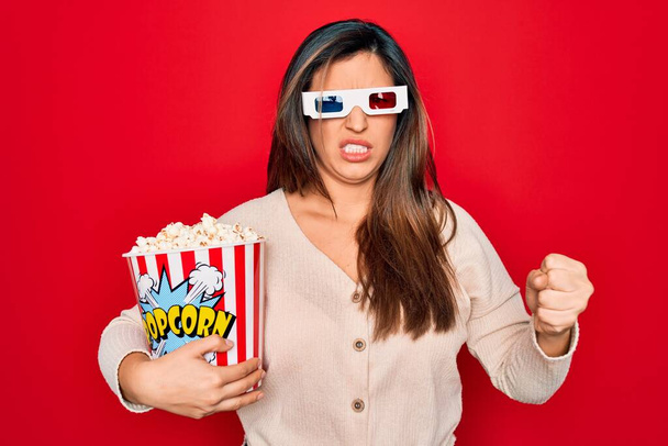 Fiatal spanyol nő visel 3D-s film szemüveg és eszik popcorn piros háttér bosszús és frusztrált kiabált harag, őrült és kiabál emelt kézzel, harag koncepció - Fotó, kép