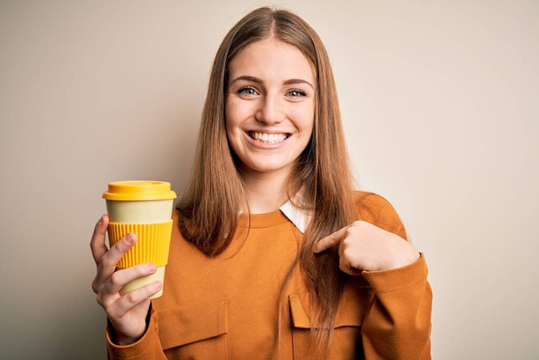 Mladý krásný ryšavý žena pití šálek kávy přes izolované bílé pozadí s překvapivou tváří ukazující prstem na sebe - Fotografie, Obrázek