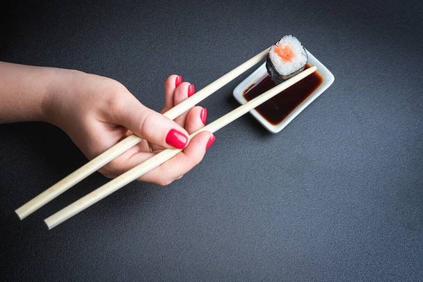 A hand holds a sushi roll with chopsticks, on a black background. Oriental cuisine. Japanese culture. - Foto, Bild