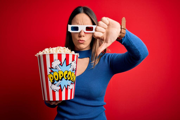 若いブロンドの女性は3Dメガネをかけて、映画館で映画を見ているポップコーンのパックを食べて、親指ダウンジェスチャーで不満と否定的な拒絶を示しています。悪い表現. - 写真・画像