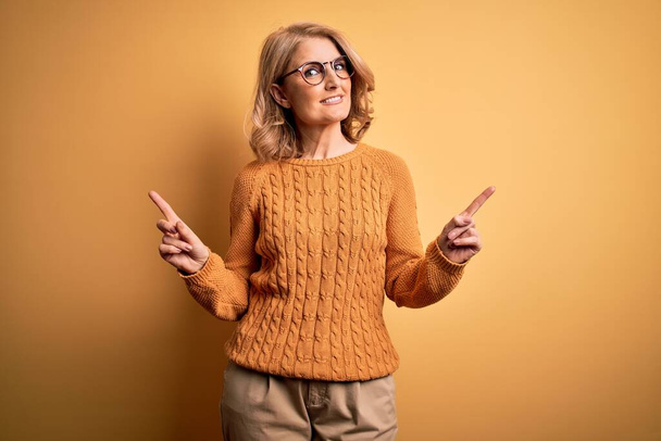 Schöne blonde Frau mittleren Alters in lässigem Pullover und Brille vor gelbem Hintergrund, die selbstbewusst lächelt und mit den Fingern in verschiedene Richtungen zeigt. Kopierfläche für Werbung - Foto, Bild