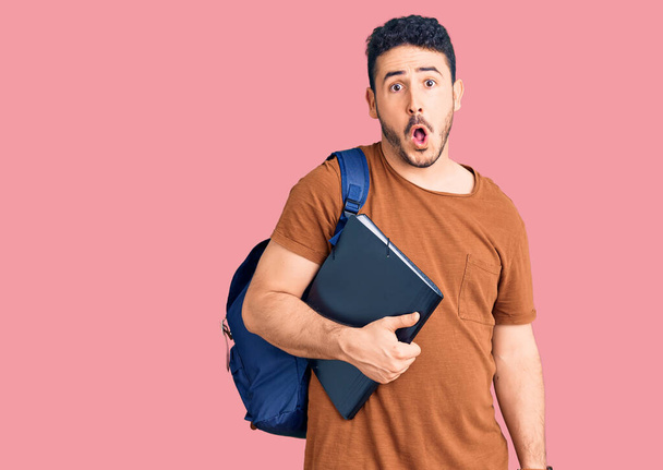Junger hispanischer Mann mit Studentenrucksack, der Ordner in der Hand hält, erschrocken und erstaunt mit offenem Mund für Überraschung, ungläubiges Gesicht  - Foto, Bild